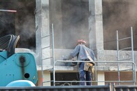 A construction worker stands with his back to the camera, a hammer drill resting on a concrete column, clouded in dust without personal protective equipment